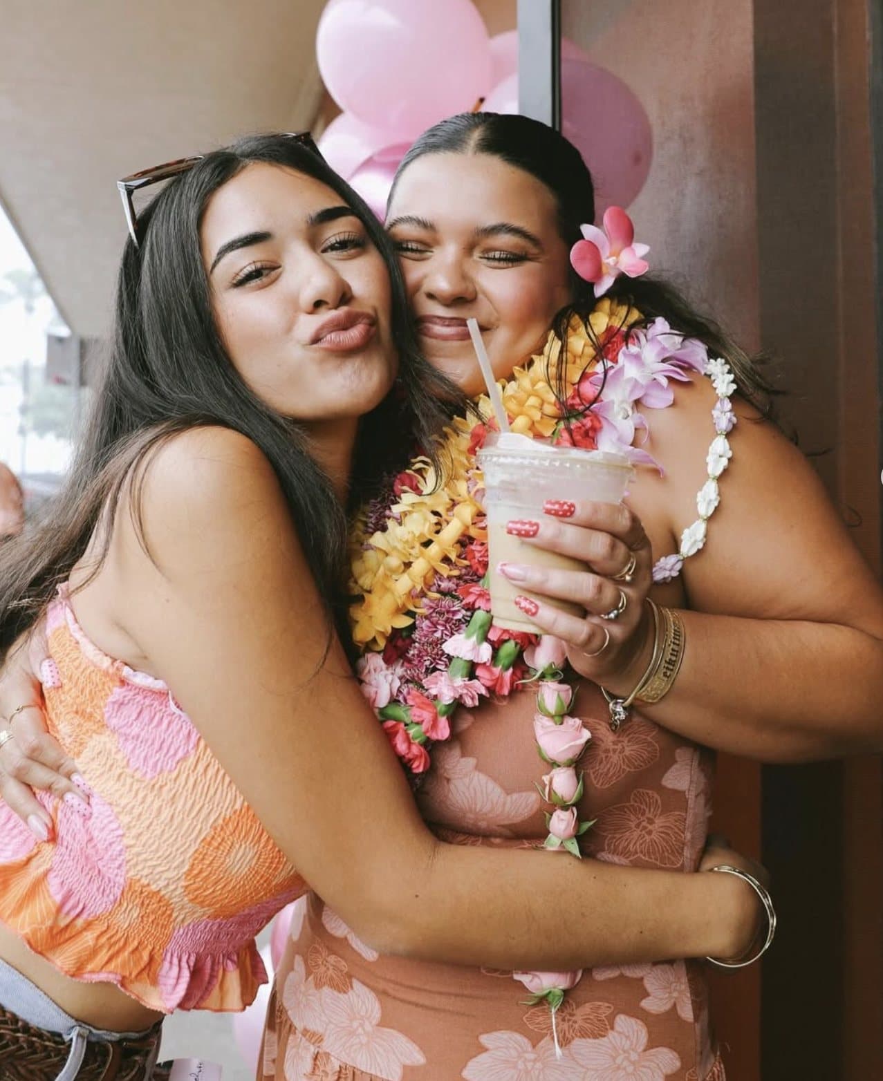 Two friends embracing at Sip n' Pretty on opening day, one holding a tropical iced drink, wearing fresh leis.