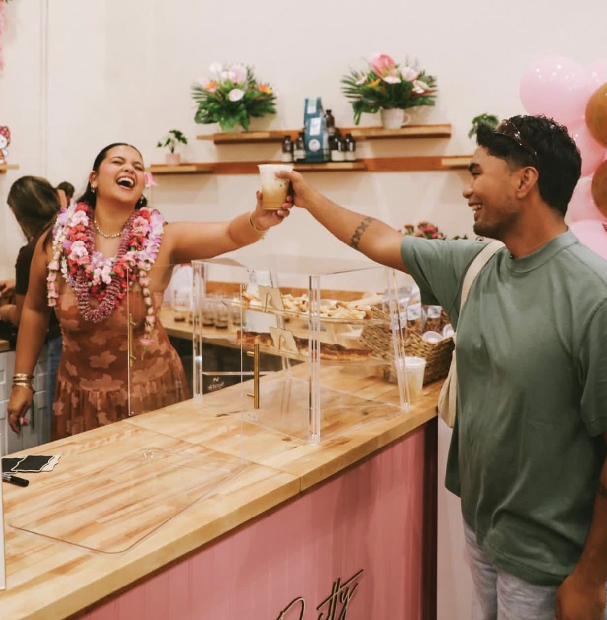 A barista and customer fist-bumping over the bar at Sip n' Pretty on opening day, donut case and pink balloons.